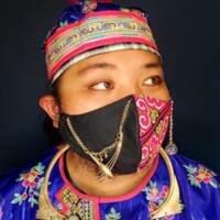 A person wearing a face mask with half of the mask designed in a traditionally female Hmong fabric pattern of pink and green, while the other half incorporates a qeej, an Hmong instrument usually played by men.