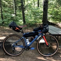 A picture of a bike step up in a wooded area with bags on it. These bags are off the back fo the seat under the top tube and on the front handlebars, used for bike packing trips and carrying clothes,food, and camping supplies.