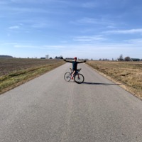 A picture of a cyclist posing in the middle of a road with barns on the horizon and farmland either side of the road