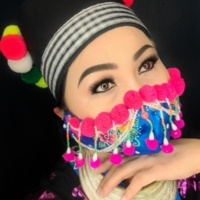 A person modeling a face mask made with traditional Hmong fabrics, designs, and embellishments.  Colors are royal blue, fuscia, yellow, and sea foam.