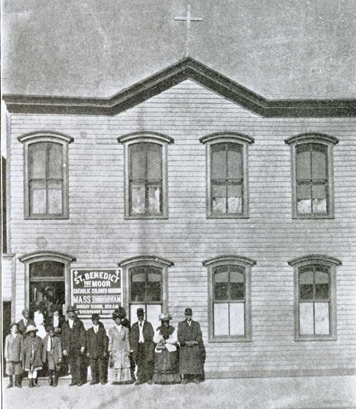 Historical photograph of the first location of St. Benedict the Moor mission. Around a dozen parishioners are standing in front of the mission.