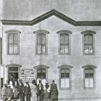 Historical photograph of the first location of St. Benedict the Moor mission. Around a dozen parishioners are standing in front of the mission.